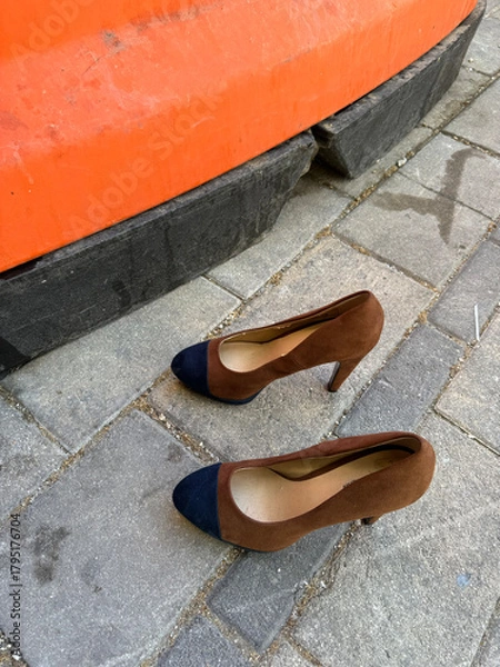 Obraz A pair of brown and navy suede high-heeled shoes rests on a cobblestone pavement next to a large orange construction barrier, suggesting a spontaneous break or abandoned footwear.