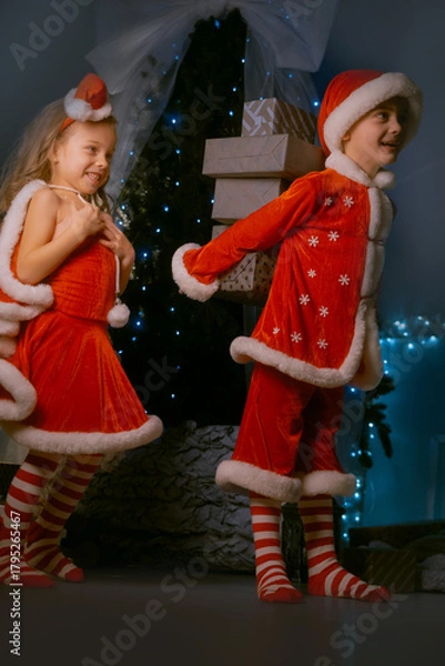 Obraz Brother and sister in red elf costumes celebrating Christmas with a large collection of presents near a cozy decorated tree.