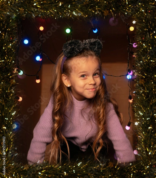 Fototapeta Happy child wearing mouse ears in a festive gift box with Christmas lights and tinsel, purple sweater, playful mood