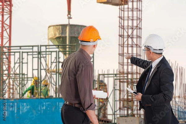 Fototapeta Two engineers in safety helmets review structural progress and coordinate development plans at a busy construction site with scaffolding and workers.