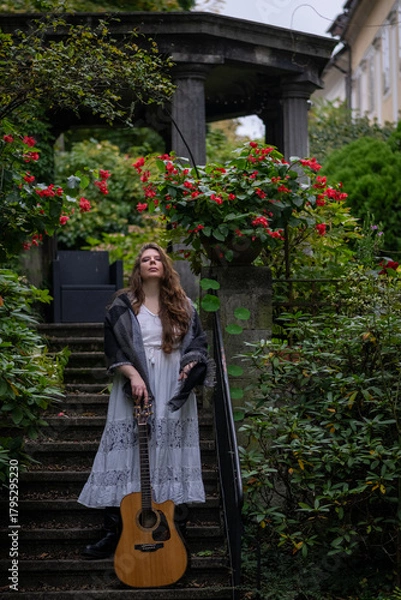Obraz Musician woman standing on stone garden stairs holding acoustic guitar with lush greenery and red flowers around