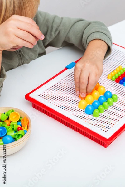 Fototapeta Child arranges colorful plastic pegs on a red mosaic board. The photo illustrates fine motor skill development, early learning, focus, and creativity through educational play. Vertical photo