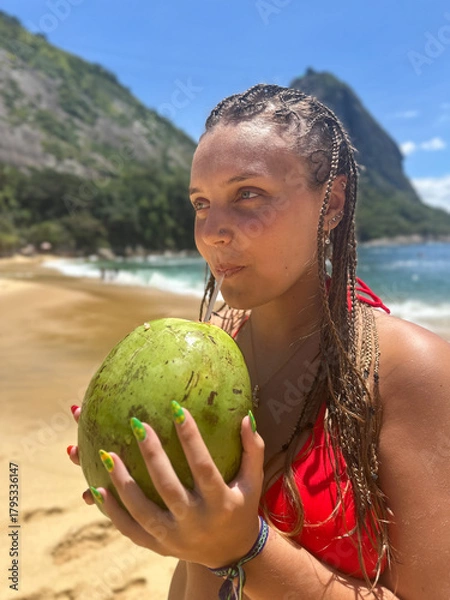 Fototapeta Young woman in a red bikini with braided hair drinking from a fresh coconut on a sunny tropical beach, relaxed and cheerful in warm natural light.