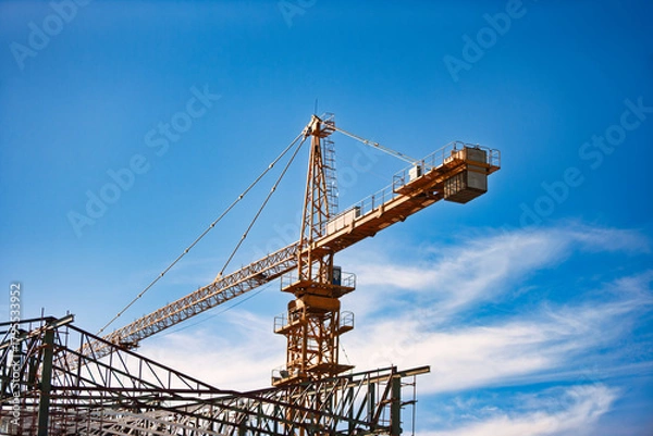 Fototapeta tower crane on a construction site, metal framework, against the blue sky with clouds