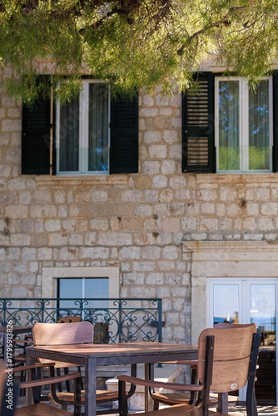 Obraz Rustic stone house with wooden table and chairs in a shaded courtyard under greenery. Concept of old town lifestyle, outdoor decor and Mediterranean charm.
