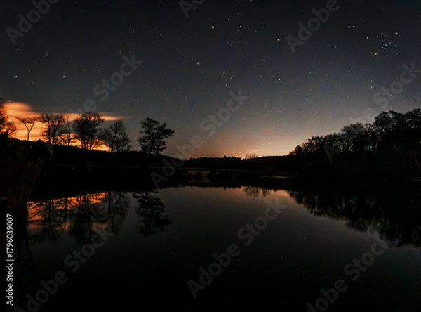 Fototapeta A star-filled night sky glows above Lake Kanawauke at Harriman State Park, where silhouetted trees and warm clouds reflect on the calm, glassy water, creating a serene, moody landscape.