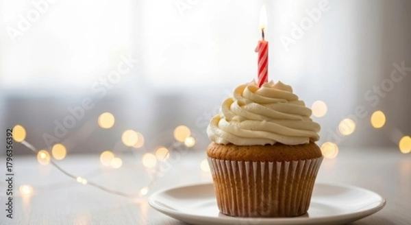 Fototapeta Single cupcake with lit candle, creamy frosting, string lights in background
