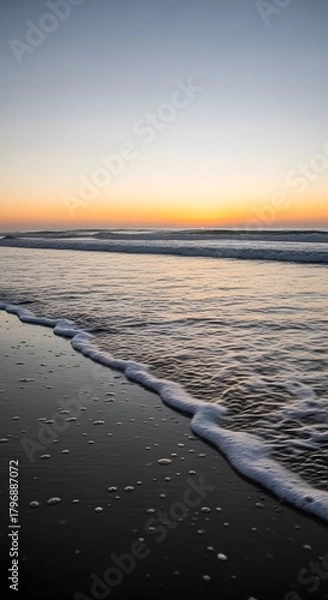 Fototapeta A serene beach scene at sunset with gentle waves washing onto the sandy shore under a colorful sky