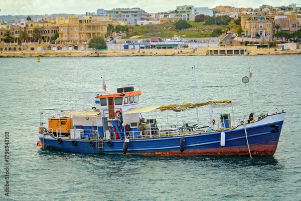 Obraz Working boat with a blue metal hull, an orange-white wheelhouse, and a yellow canopy floating on calm water near the coast with a scenic urban background.