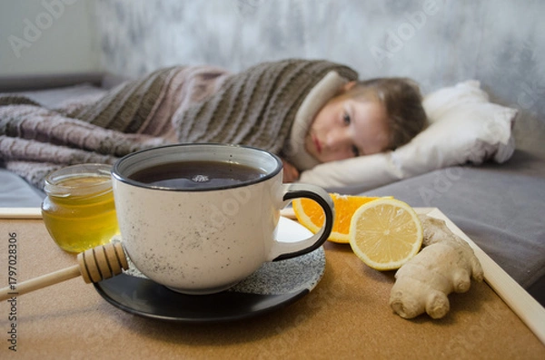 Fototapeta A little girl lies in bed drinking tea with lemon. Concepts of illness, cold, and acute respiratory viral infection.	