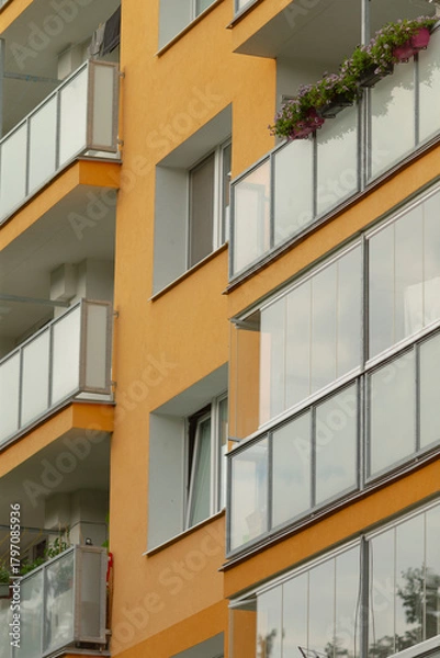 Fototapeta A close-up view of a modern apartment building featuring a vibrant orange facade and balconies adorned with flowers, showcasing contemporary architecture.