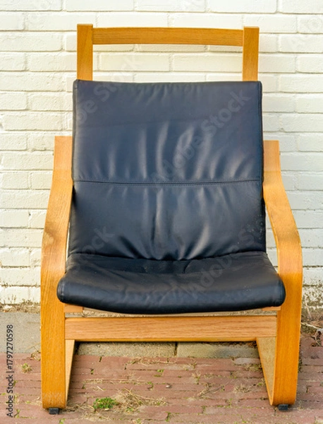 Obraz wooden armchair with a black leather cushion stands outside in front of a cream-colored painted wall