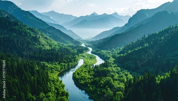 Obraz Serpentine River Winds Through Lush Green Mountain Valley Under Soft Blue Sky Hazy Distant Peaks Bathed in Sunlight