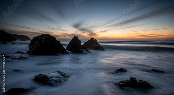 Fototapeta A serene coastal landscape captured with long exposure, showing the dynamic motion of streaking clouds and misty sea around silhouetted rocks at sunset