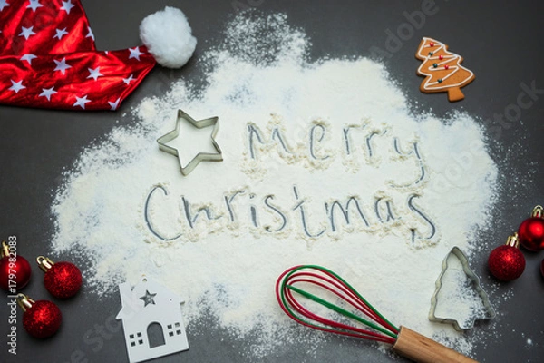 Fototapeta Flour surface with a handwritten Merry Christmas message, festive ornaments, cookie cutters, and a Santa hat creates a warm seasonal mood. Ideal for holiday advertising, baking themes, and designs.