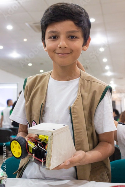 Fototapeta Portrait of a Hispanic boy smiling and looking at the camera with a robot in his hands inside an auditorium in Neiva, Huila, Colombia. Concept of science and robotics