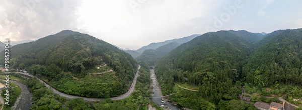 Obraz Panoramic view of a winding road through a lush, green valley flanked by mountains. A river flows alongside the road, reflecting the cloudy sky. A small building is visible on the right.