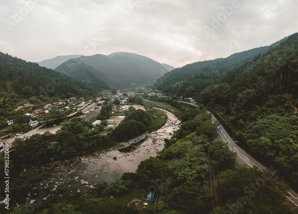 Obraz Aerial view of a winding river valley with lush green forests and a small town nestled along the banks. A road follows the river's course through the mountains. Overcast sky adds a moody atmosphere.