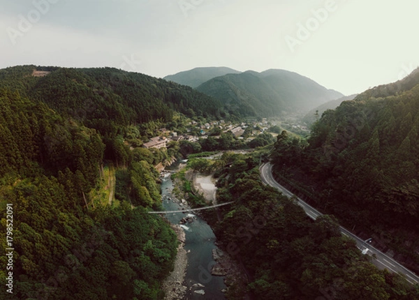 Obraz Aerial view of a mountainous valley with a river, road, and small town. Lush green forests dominate the landscape under a cloudy sky. Serene and picturesque.