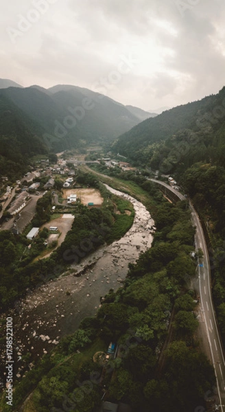 Obraz Aerial view of a river winding through a lush, mountainous valley. A road follows the river's course, passing through a small town. Overcast sky adds to the moody atmosphere.