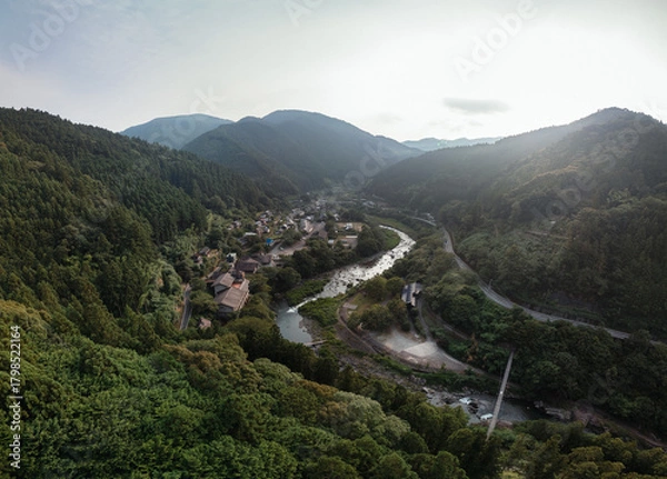 Obraz A scenic aerial view of a village nestled in a valley alongside a winding river, surrounded by lush green mountains and forests. The scene evokes a sense of tranquility and natural beauty.