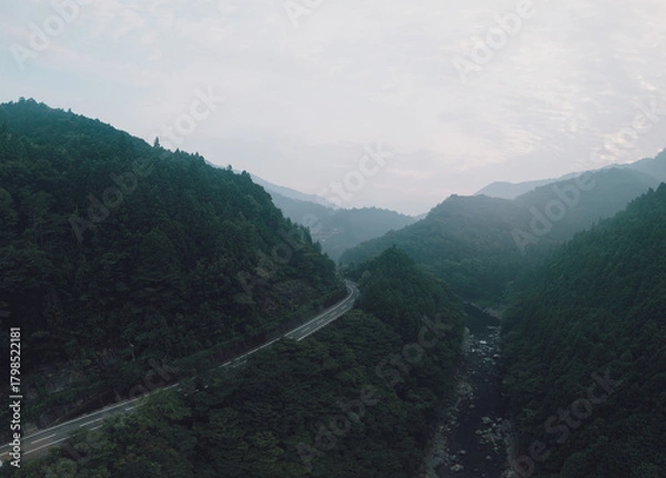 Obraz A scenic aerial view of a winding road through a lush, mountainous landscape shrouded in mist. The road cuts through dense green forests and a river valley.