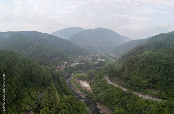 Obraz Aerial view of a lush valley with a winding river, surrounded by dense forests and mountains. A road snakes through the landscape, leading to a small town nestled among the trees. 