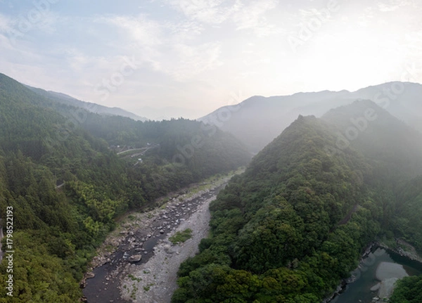 Obraz Aerial view of a lush, mountainous valley with a winding river and dense forest. Mist hangs in the air, creating a serene and atmospheric landscape.