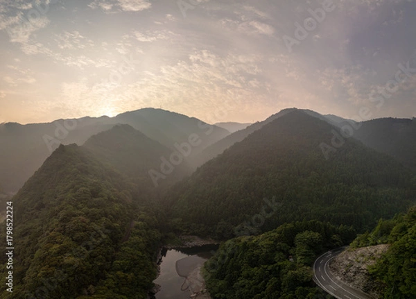 Obraz A stunning aerial view of a mountain range at sunrise, with a winding road cutting through a lush valley and a river flowing through it. The sky is filled with dramatic clouds.
