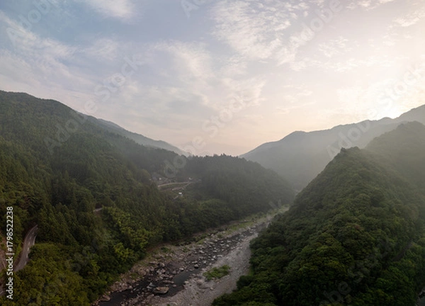Obraz A stunning aerial view of a lush, green valley with a winding river and towering mountains under a cloudy sky at sunrise.