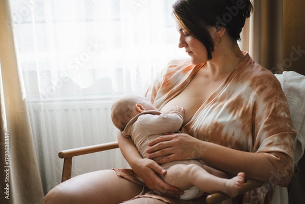 Obraz A mother sits in a wooden chair, gently breastfeeding her baby in a warm and softly lit room. Sunlight filters through the curtains, creating a peaceful atmosphere.