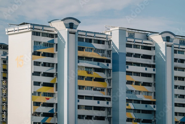 Obraz A colorful HDB block in Singapore with a distinctive geometric design. The building features a mix of white, yellow, and blue, with a prominent "15" on one side. The sky is blue with some clouds.