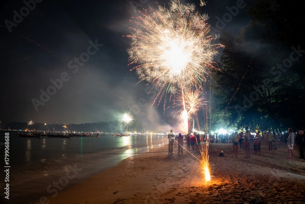 Fototapeta Long exposure shot capturing large white and gold fireworks exploding above a sandy beach with sparks rising from the ground and dark silhouettes of people watching