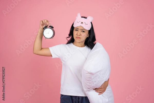 Fototapeta A tired and sleepy Asian woman holds an alarm clock and a pillow, standing isolated on a pink background.