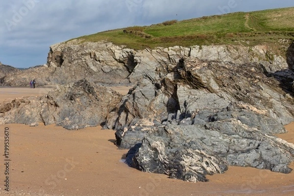 Fototapeta grey rocks on a sandy beach at the foot of a green hillside under a cloudy sky in Polly Joke - Cornwall - UK