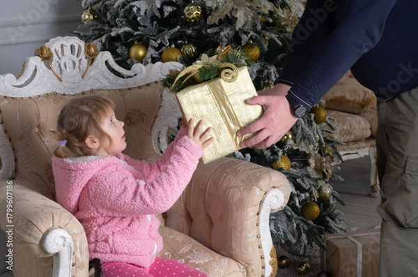 Fototapeta a little girl in a pink sweater is sitting in a chair and being handed a large box wrapped in Christmas paper