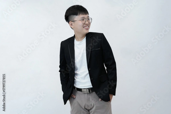 Fototapeta A cheerful and relaxed young Asian professional or student standing with hands in their pockets, smiling comfortably against a clean white studio background