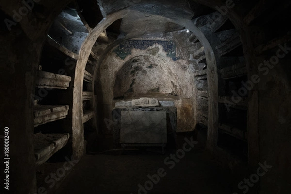 Fototapeta Dimly lit ancient catacomb chamber with stone shelves, altar and fresco fragments - atmospheric underground crypt in Naples, Italy