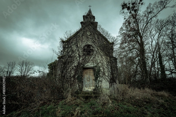 Fototapeta Vine-covered abandoned chapel with weathered wooden door and round window beneath a moody, overcast sky