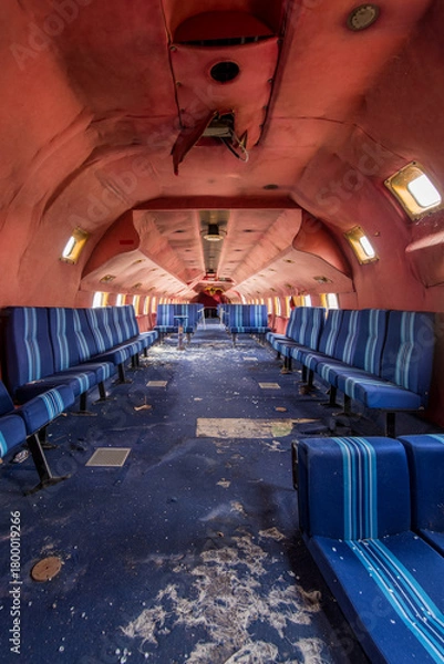 Fototapeta Derelict airplane cabin with blue striped seats and peeling red ceiling - abandoned aircraft interior with a long symmetrical aisle and scattered debris