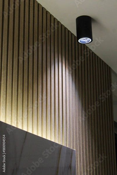 Obraz Close-up of a sleek black wall light fixture illuminating a white wall in a minimalist interior, creating elegant contrast and soft ambient lighting.