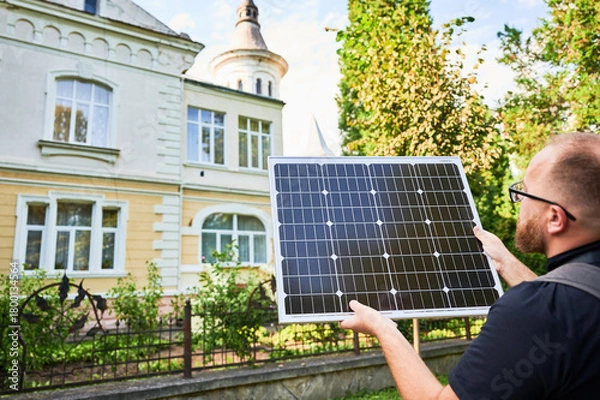Fototapeta Man holding photovoltaic solar panel in front of historical building. Concept of integration of sustainable renewable energy sources into architecture.
