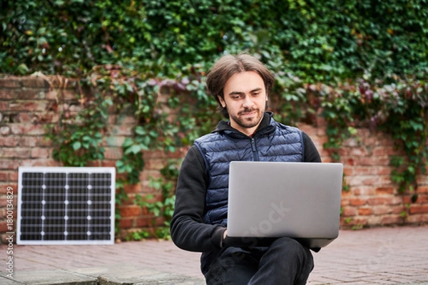 Fototapeta Man sits outdoors, working on laptop against backdrop of ivy-covered brick walls. Solar panel rests nearby, highlighting sustainable, eco-friendly workspace in serene, natural urban environment.