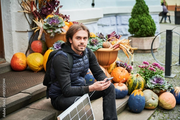 Fototapeta Handsome man using smartphone connected to photovoltaic solar panel. Integration of sustainable renewable energy into everyday life, demonstrating practical use of solar power for charging devices.