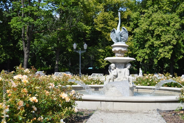 Fototapeta Fountain Among Blooming Roses. A decorative stone fountain with sculpted cherub-like figures pours clear streams of water into a circular basin, surrounded by blooming roses and greenery