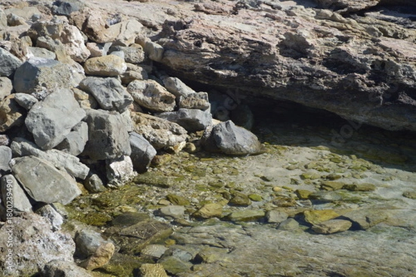 Fototapeta Close-up of rocky shore with clear tidal pool and pebbles under bright sunlight, peaceful coastal nature, copy space
