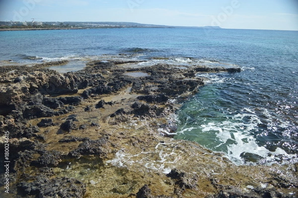 Fototapeta Rocky coastline with clear water and gentle waves under bright blue sky, picturesque coastal nature, copy space