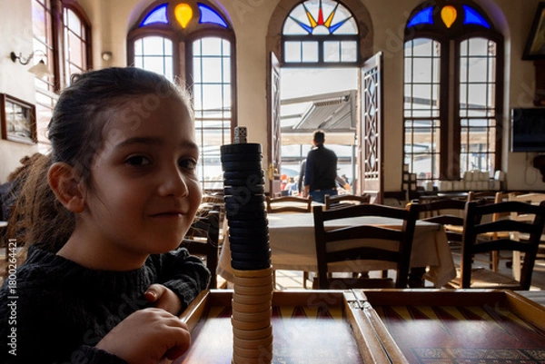 Fototapeta Young girl sitting in a traditional café playing with stacked backgammon pieces, with sunlight shining through stained-glass windows and people in the background, capturing a warm everyday scene.