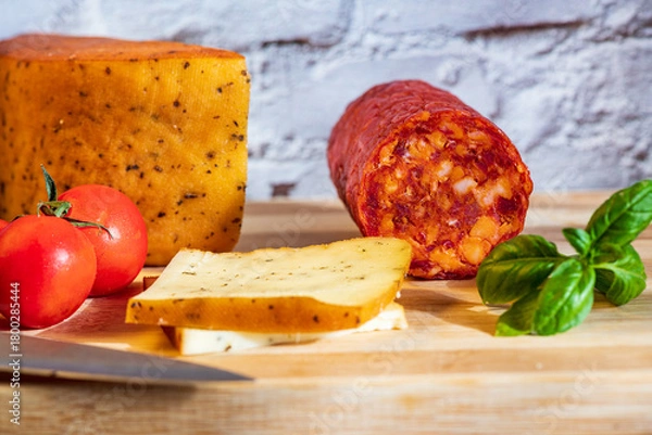 Fototapeta Sliced kulen and semi-hard cheese arranged neatly on a plate, garnished with fresh basil leaves. The vibrant colors of the red cured sausage, creamy-yellow cheese, and green basil create a visually ap