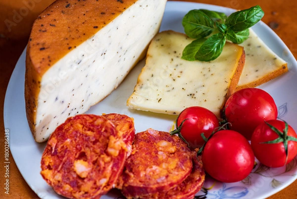Fototapeta Sliced kulen and semi-hard cheese arranged neatly on a plate, garnished with fresh basil leaves. The vibrant colors of the red cured sausage, creamy-yellow cheese, and green basil create a visually ap